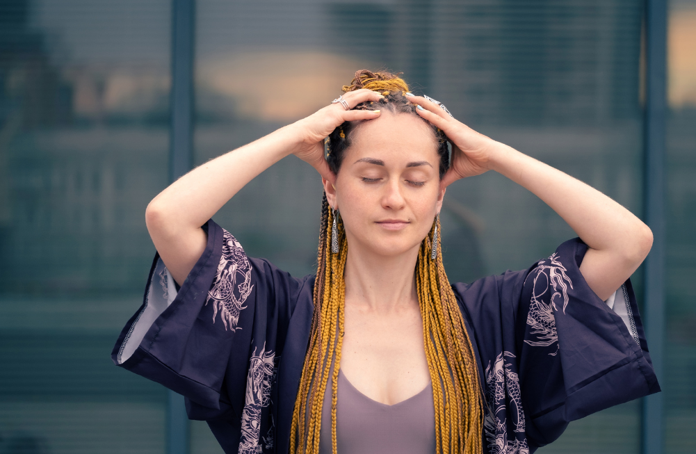 a woman with a long hair wearing a kimono sleeve boho outfit covering her head with both hands closed eyes and relaxed.